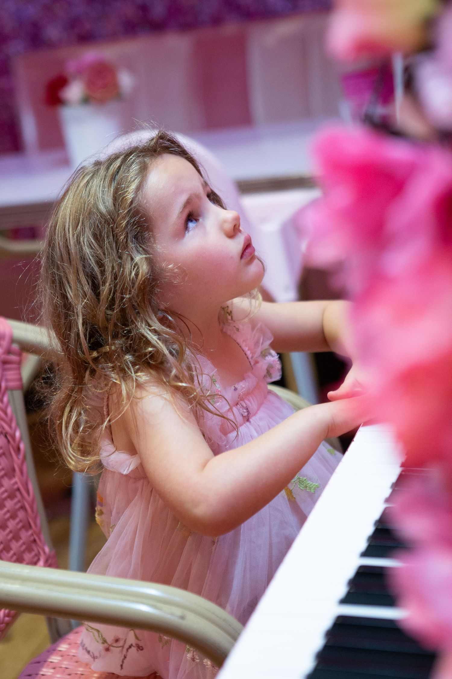 Young girl in pink dress playing piano keys while looking up with curiosity in a pink decorated room.
