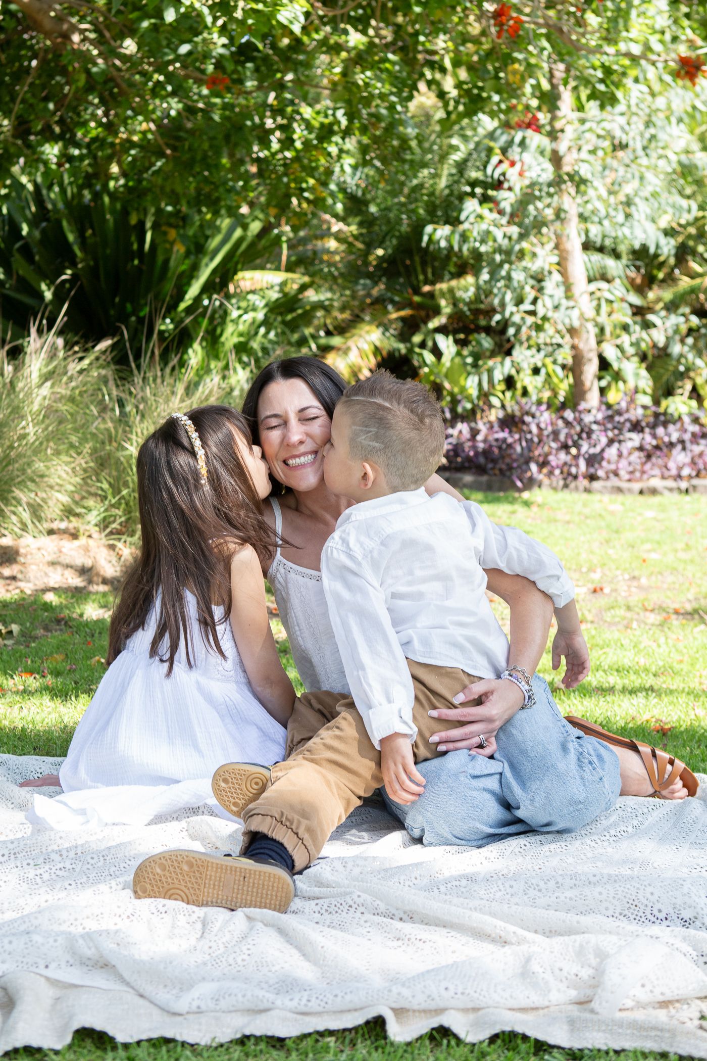 A mother sits on a blanket in a park, smiling as her two young children kiss her on the cheeks.