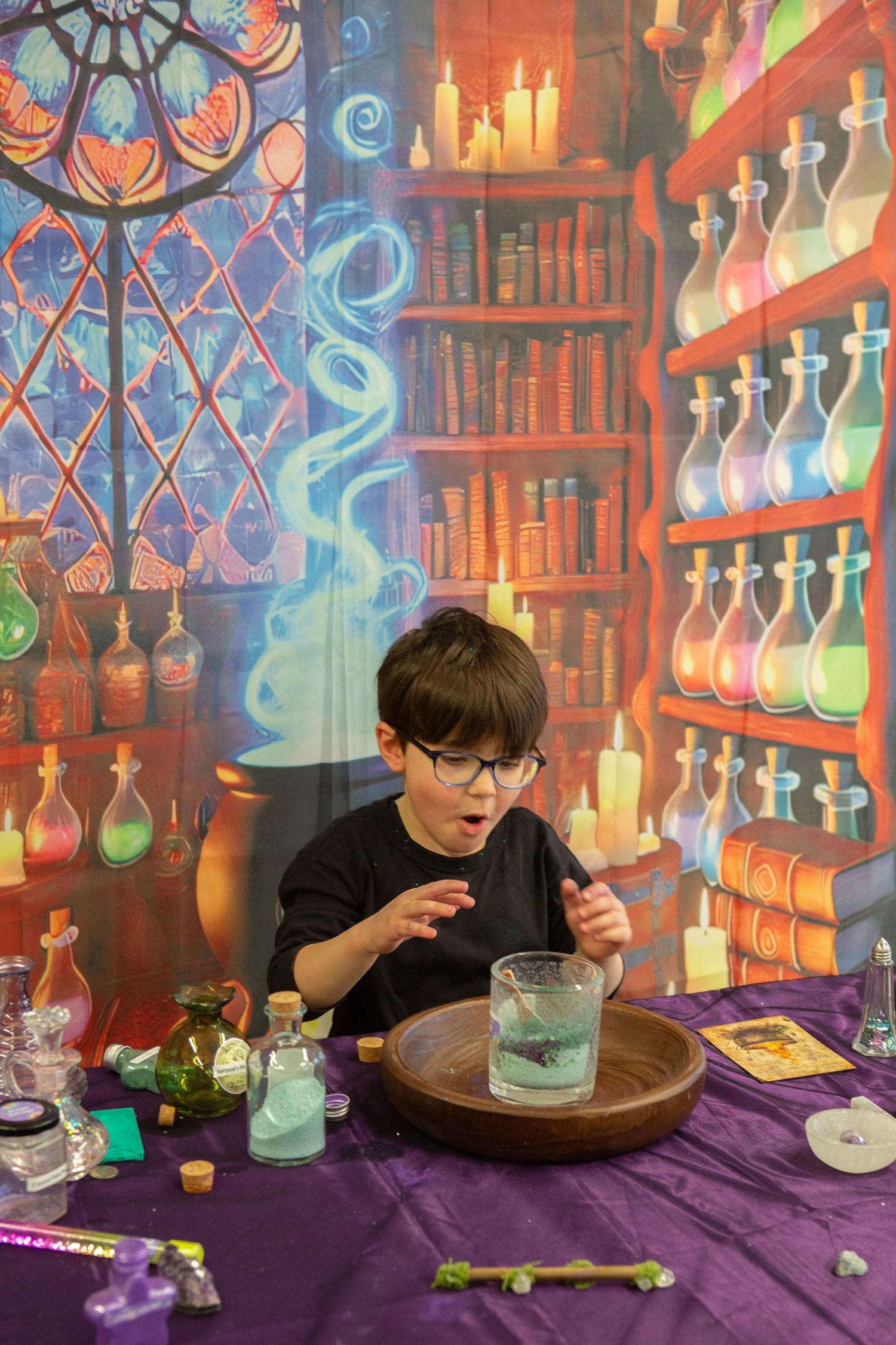 Young boy in glasses doing a magic potion experiment at a table with a fantasy wizard laboratory backdrop.
