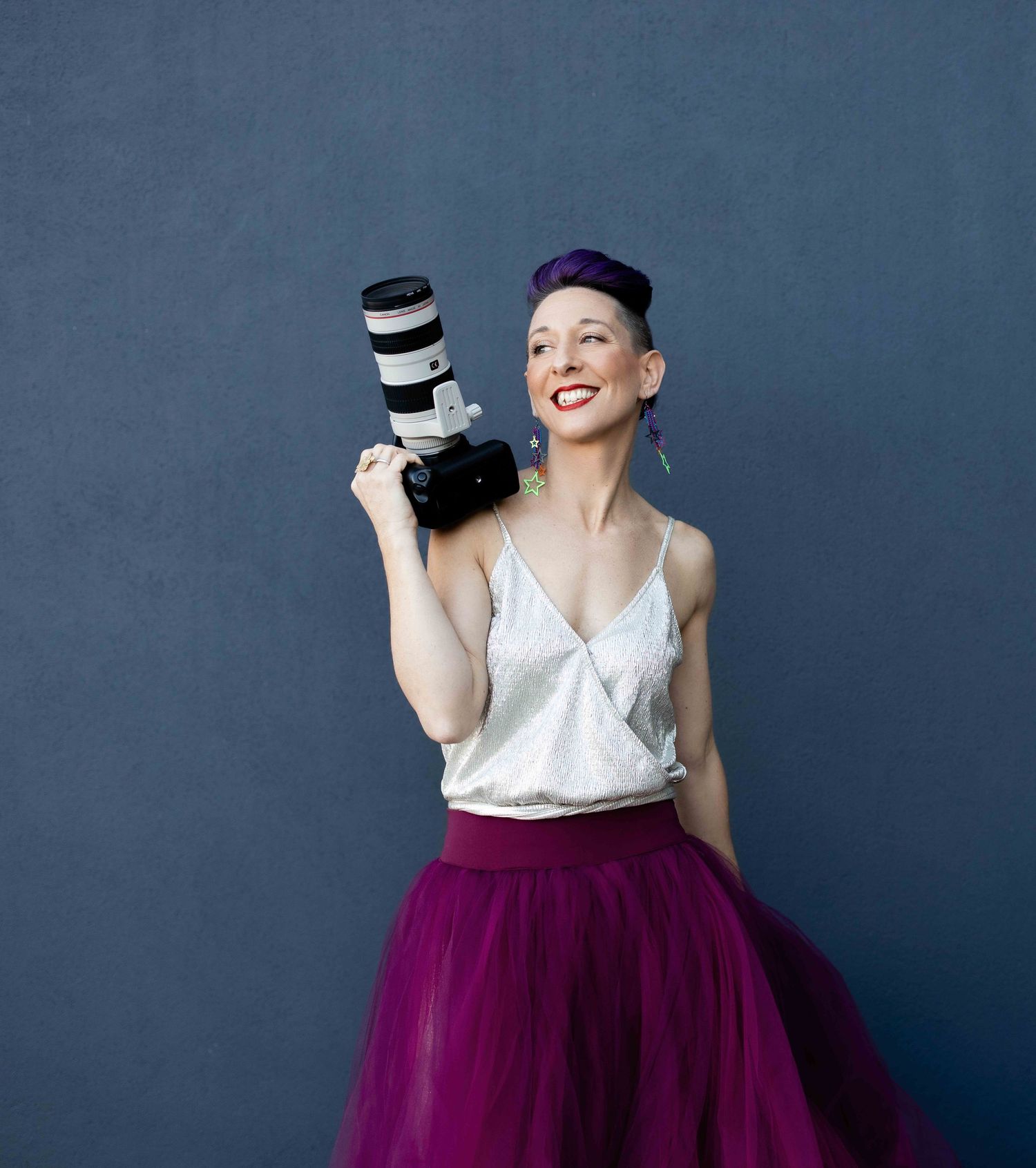 Photographer in purple cap and tulle skirt holding professional camera against blue background.