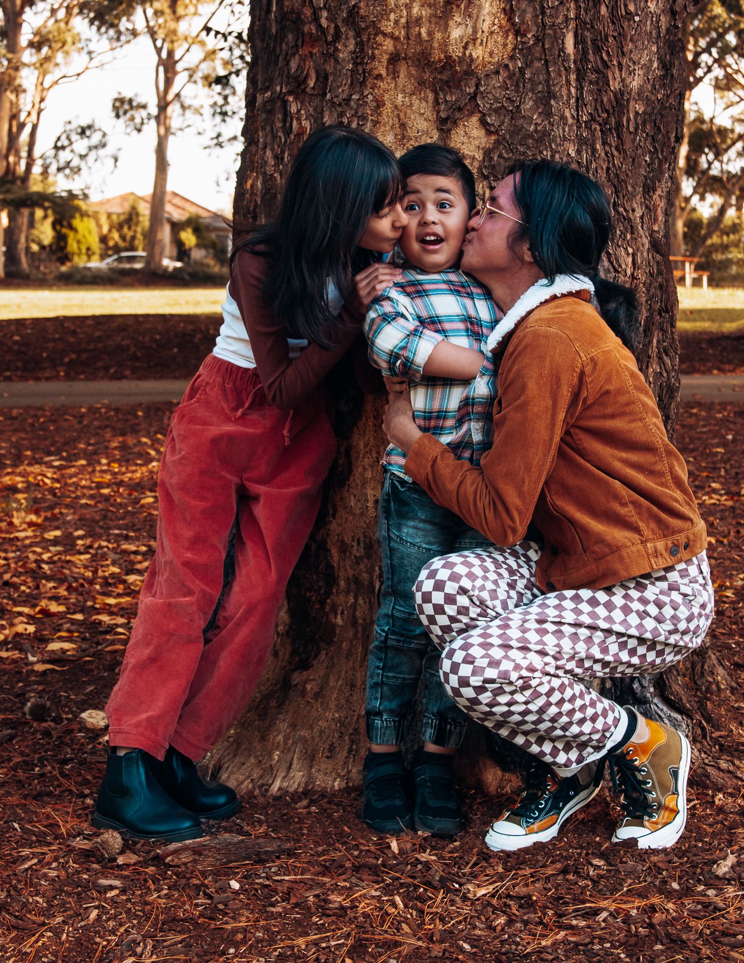 Two women kissing a young boy on the cheeks beside a tree in an autumn park.