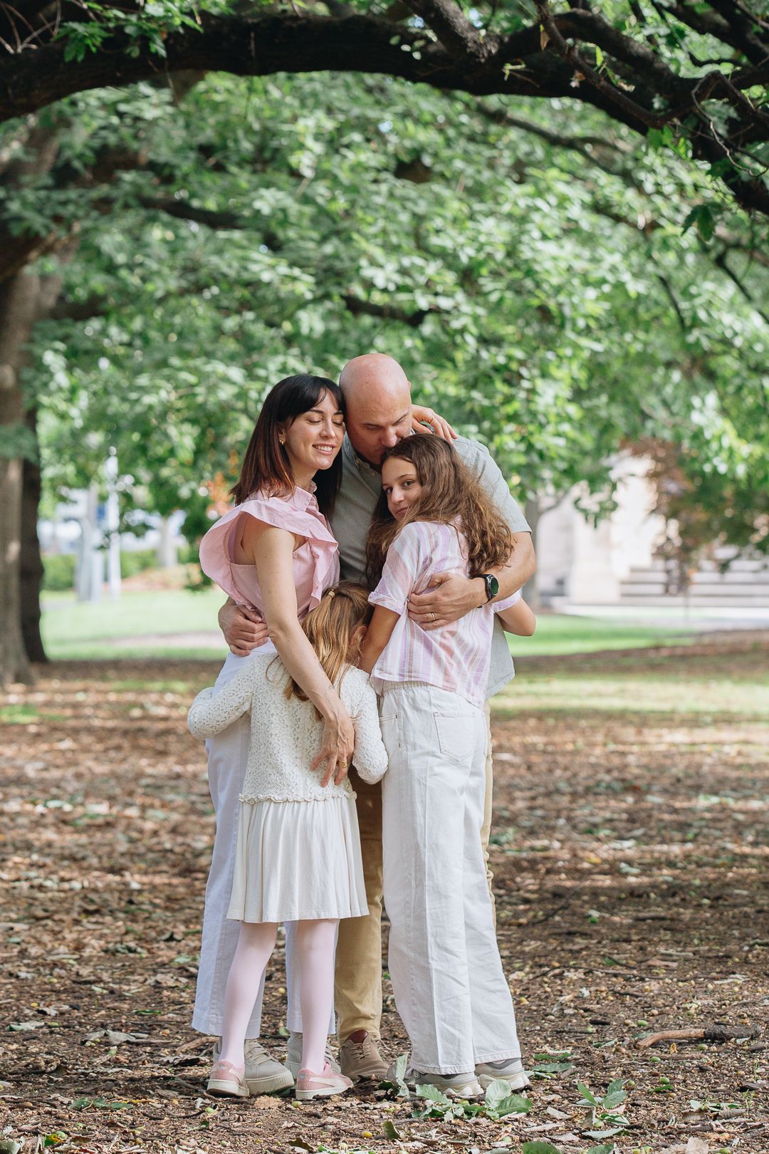 Happy family of four sharing a warm group hug in a lush green park, parents embracing two daughters outdoors.