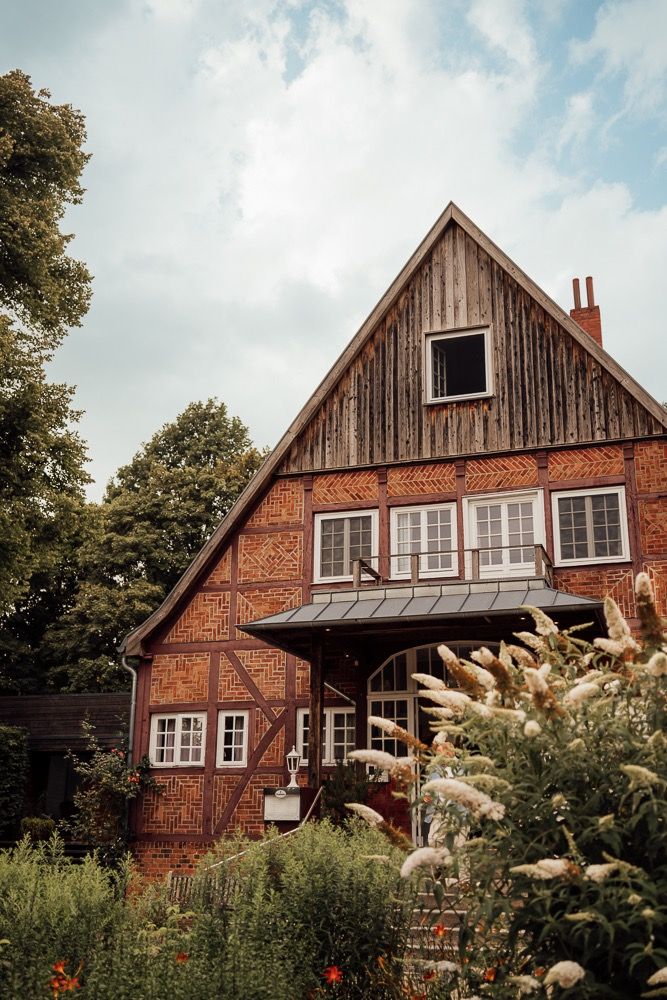 Rustic red brick and wood farmhouse with white trim windows photographed from multiple angles in a lush garden setting.