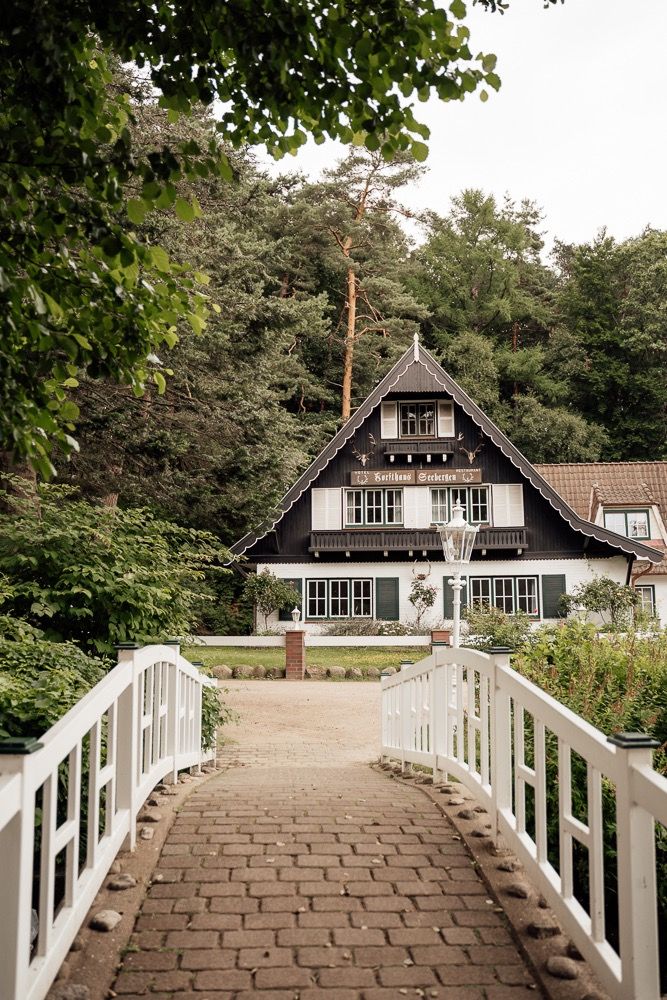White wooden bridge leads to charming black and white cottage houses nestled among trees in a tranquil setting.