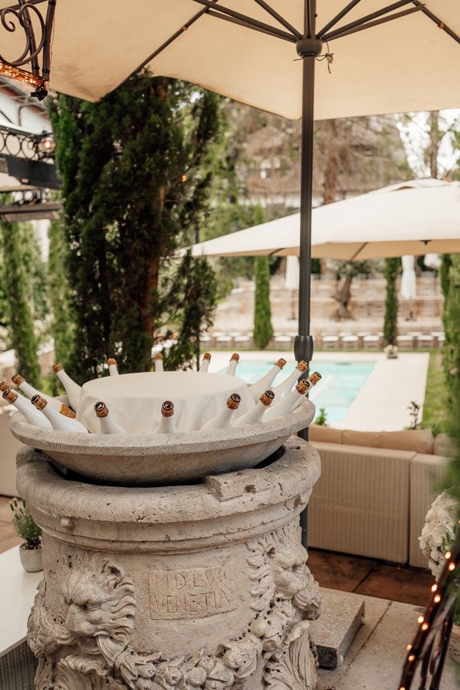 Ornate stone champagne bucket with bottles chilling on an elegant patio with pool and umbrellas in background.