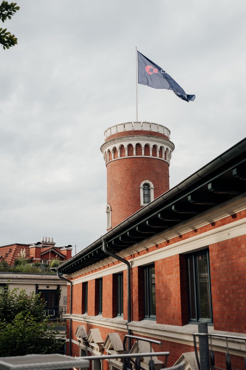 Red brick building with cylindrical tower flying a blue flag against cloudy sky.