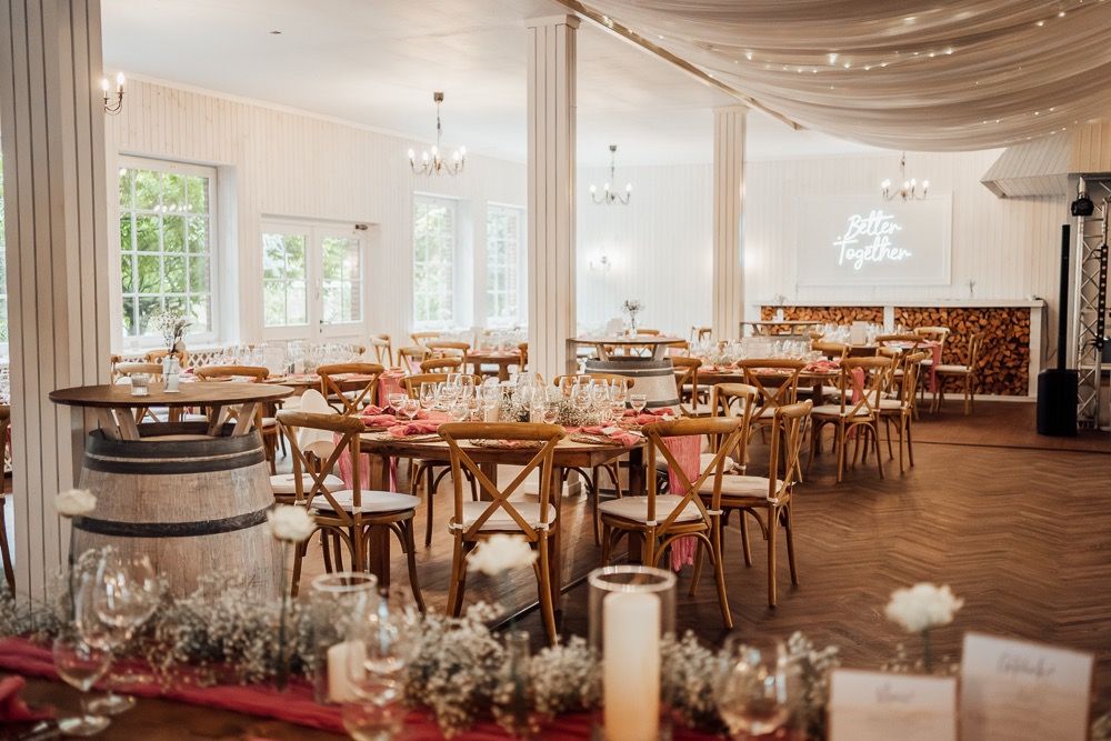 Spacious white banquet hall decorated with twinkle lights, fabric draping, and wooden cross-back chairs for a wedding.