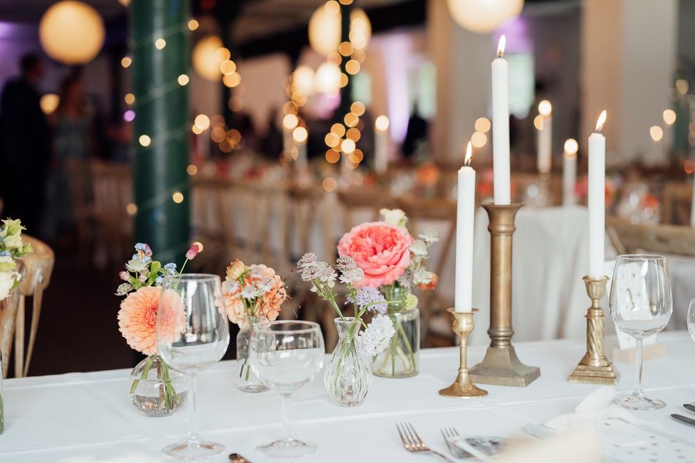 Elegant wedding table setting with pink roses, tall white candles in brass holders, and wine glasses on white tablecloth.