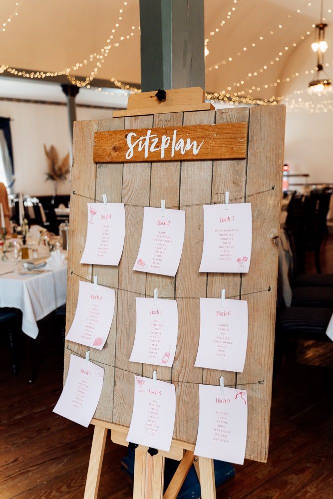 Wedding seating chart display with white cards pinned on rustic wooden boards under twinkle lights at reception venue.