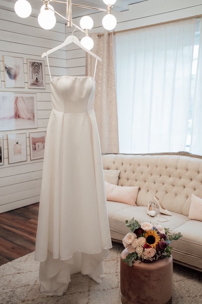 A white wedding dress hangs elegantly in a bridal suite with tufted furniture and floral arrangements.