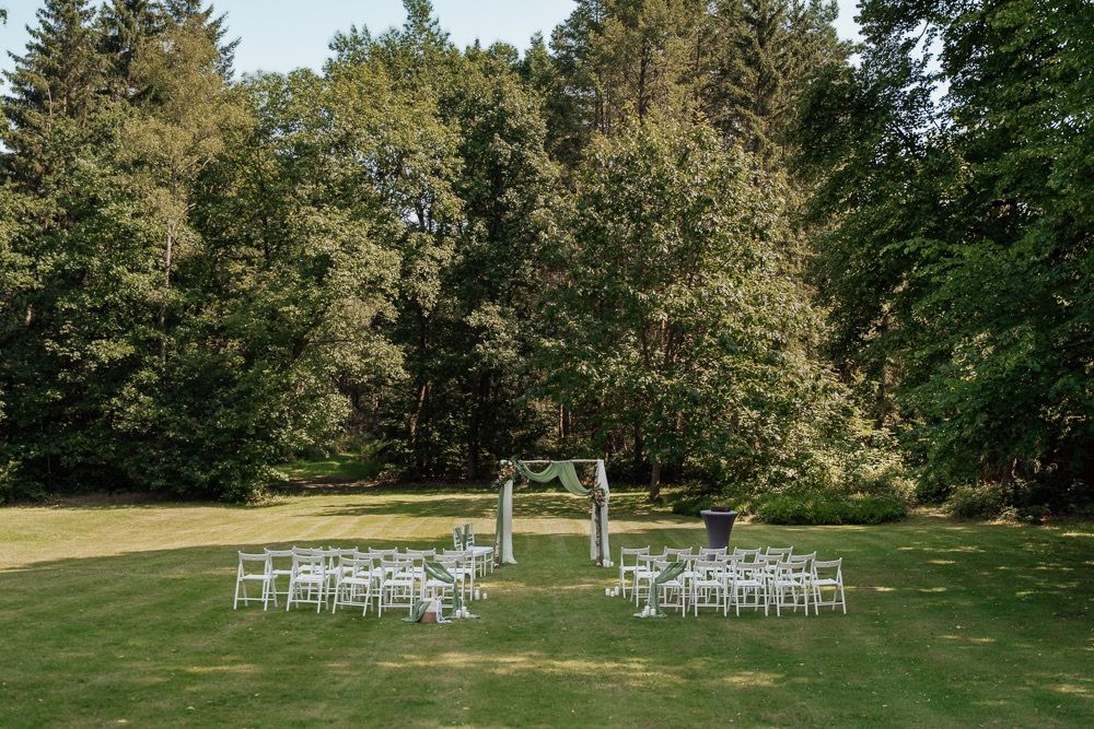 White chairs arranged for an outdoor wedding ceremony with a wooden arch on a green lawn surrounded by tall evergreen trees.