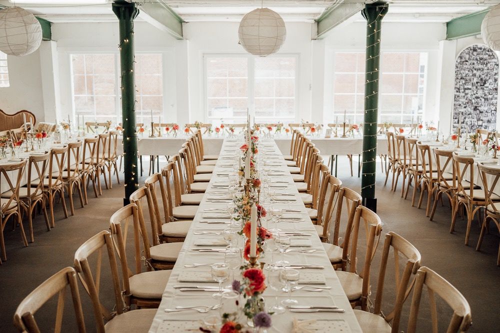 Long banquet tables with white tablecloths and wooden chairs set up in an elegant reception hall with natural lighting.