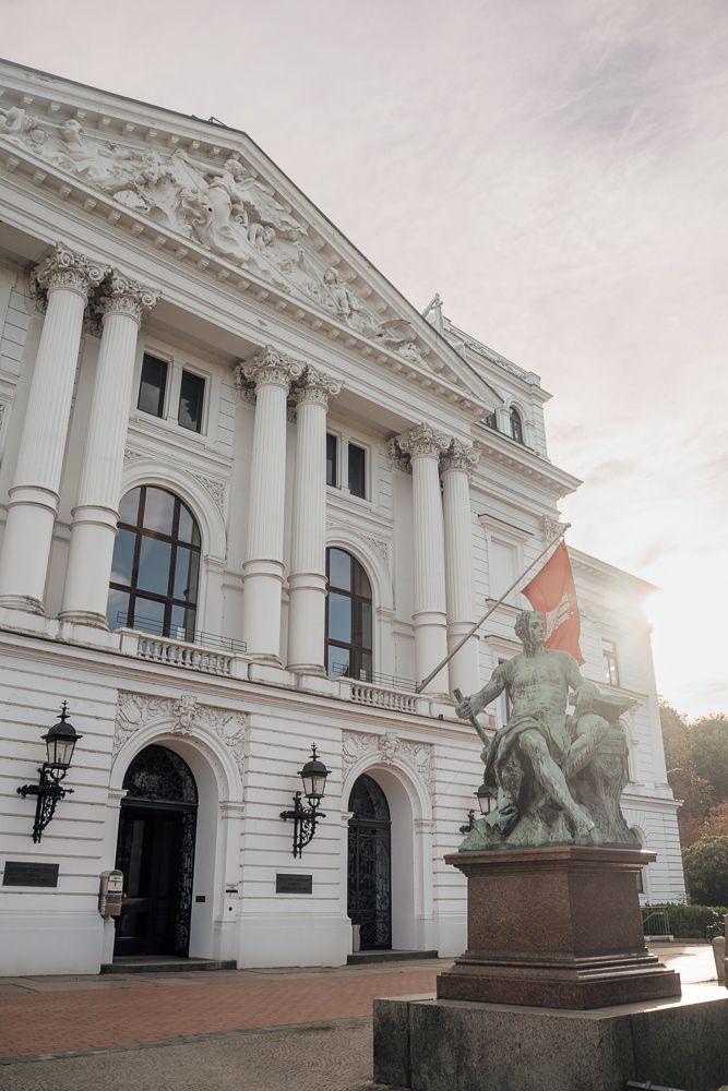 Ornate white neoclassical building facade with tall columns and equestrian statue at entrance in soft sunlight.