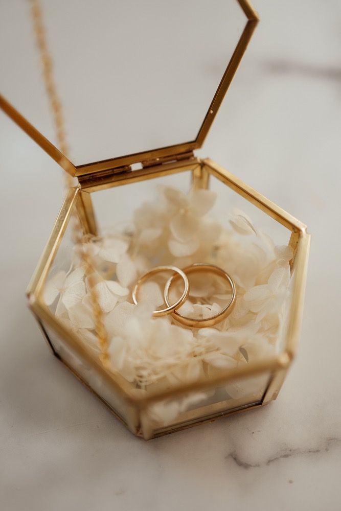 Two gold wedding rings rest in a hexagonal glass box filled with delicate white petals on a marble surface.