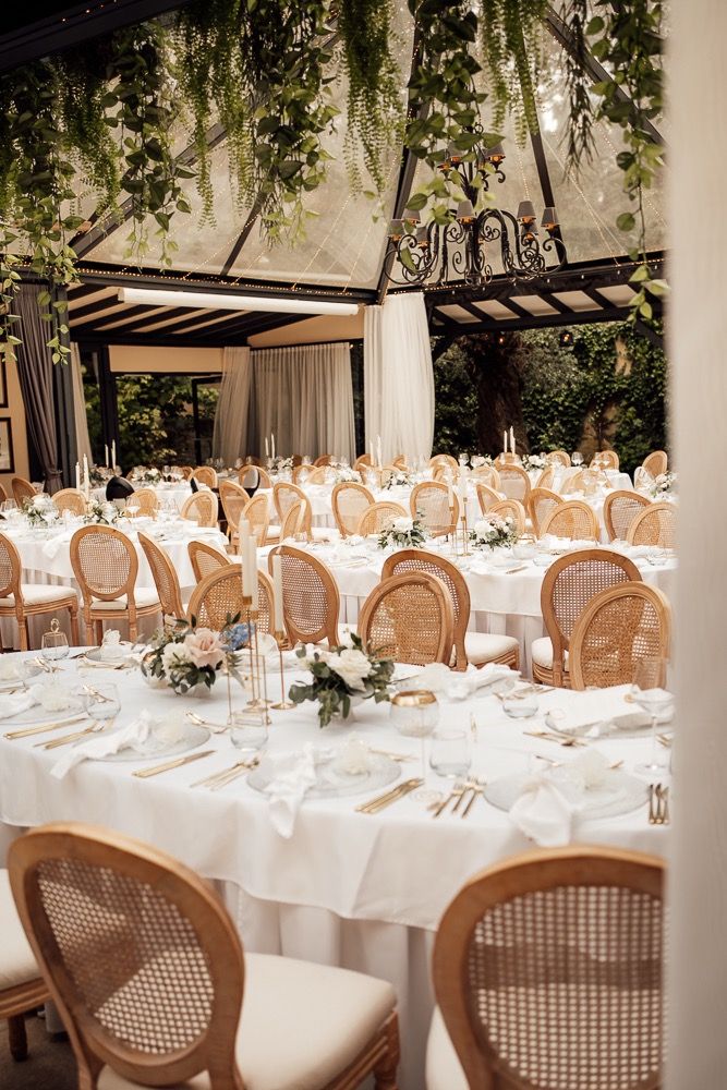 Elegant wedding reception tables set with white linens and cane-back chairs in an outdoor covered venue with natural lighting.
