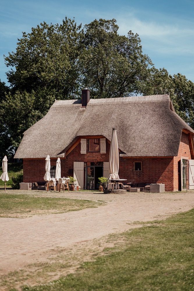 Traditional thatched-roof brick cottage with outdoor seating and umbrellas in a rural countryside setting.