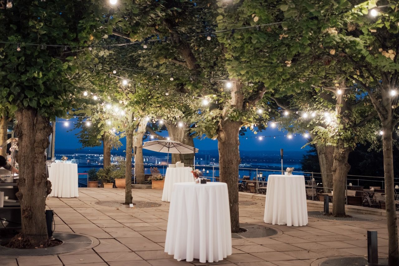 Elegant outdoor cocktail reception with white draped tables under string lights and lush trees at twilight.