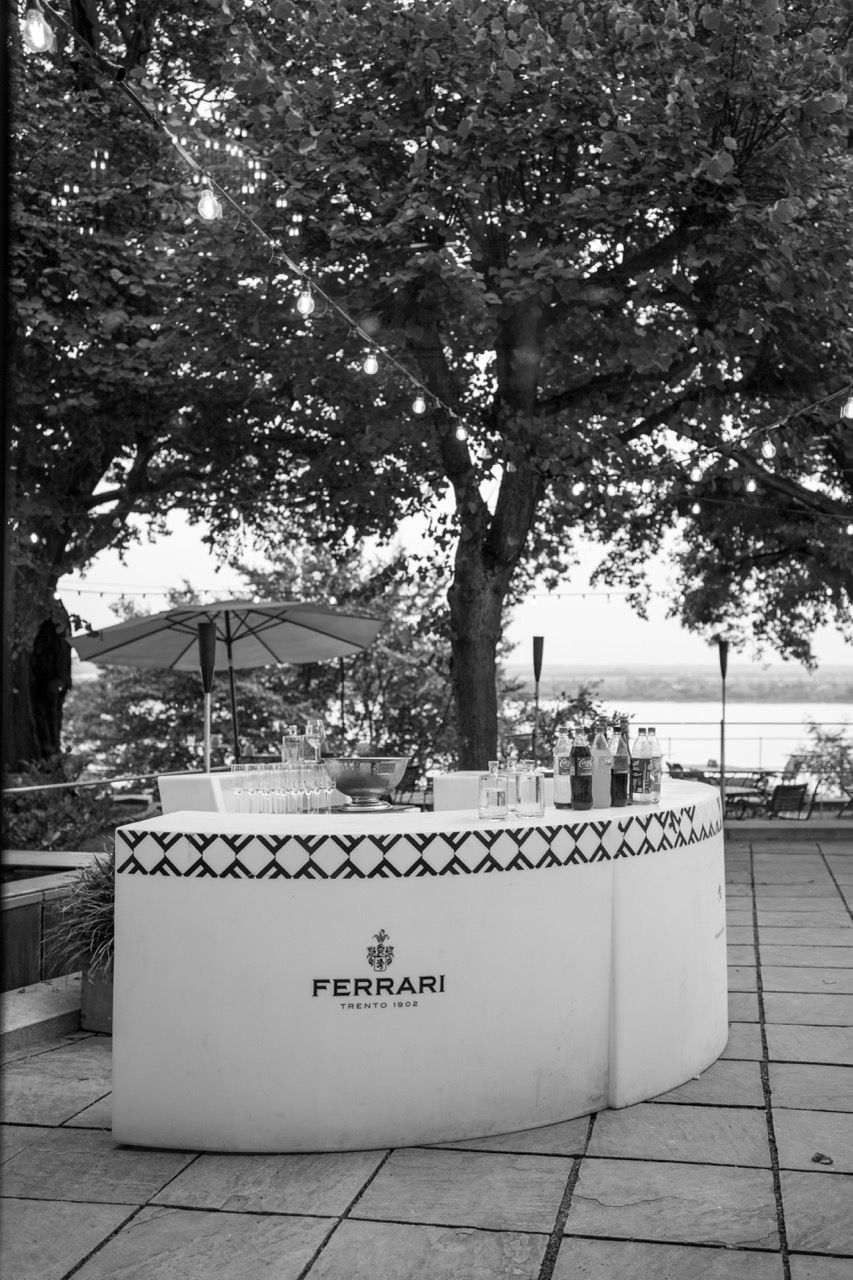 Black and white photo of an elegant outdoor Ferrari Trento sparkling wine bar surrounded by lush trees near a waterfront.
