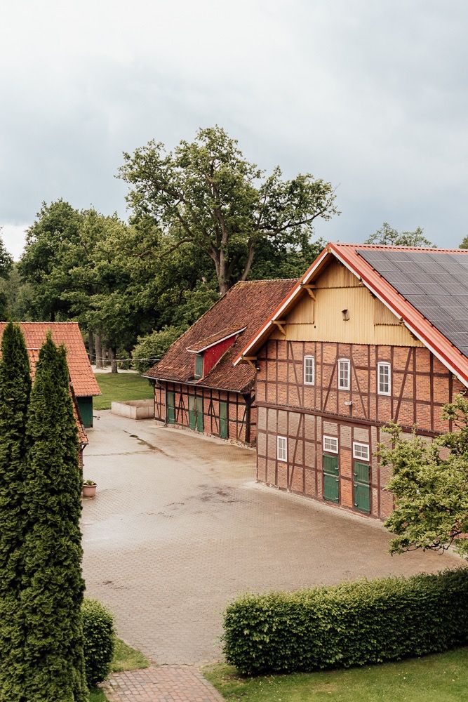 Row of traditional half-timbered barns with red roofs and beige walls along a paved courtyard in rural Germany.