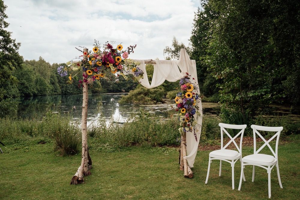 White chairs and floral arrangements create an elegant outdoor wedding ceremony setup in a lush garden setting.
