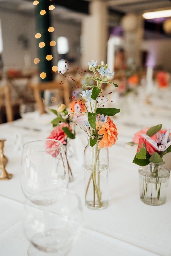 Small glass vases with delicate pink and coral flowers create an elegant table setting with white linens and twinkling lights.