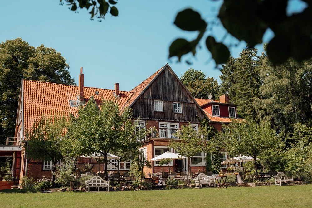 Rustic wooden lodge with red roof nestled among trees features outdoor seating area on sunny summer day.