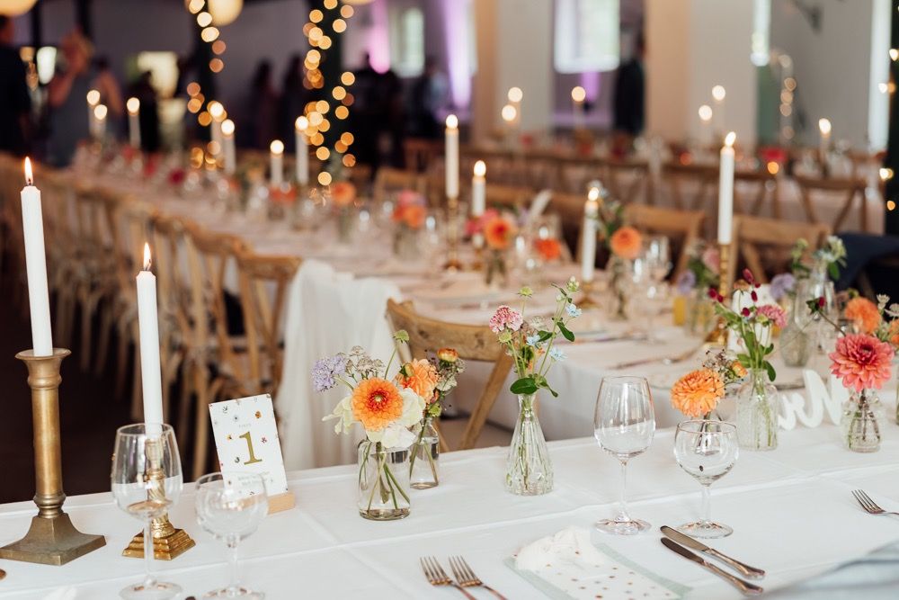 Elegant wedding reception tables decorated with floral centerpieces, candles and place settings in a bright indoor venue.
