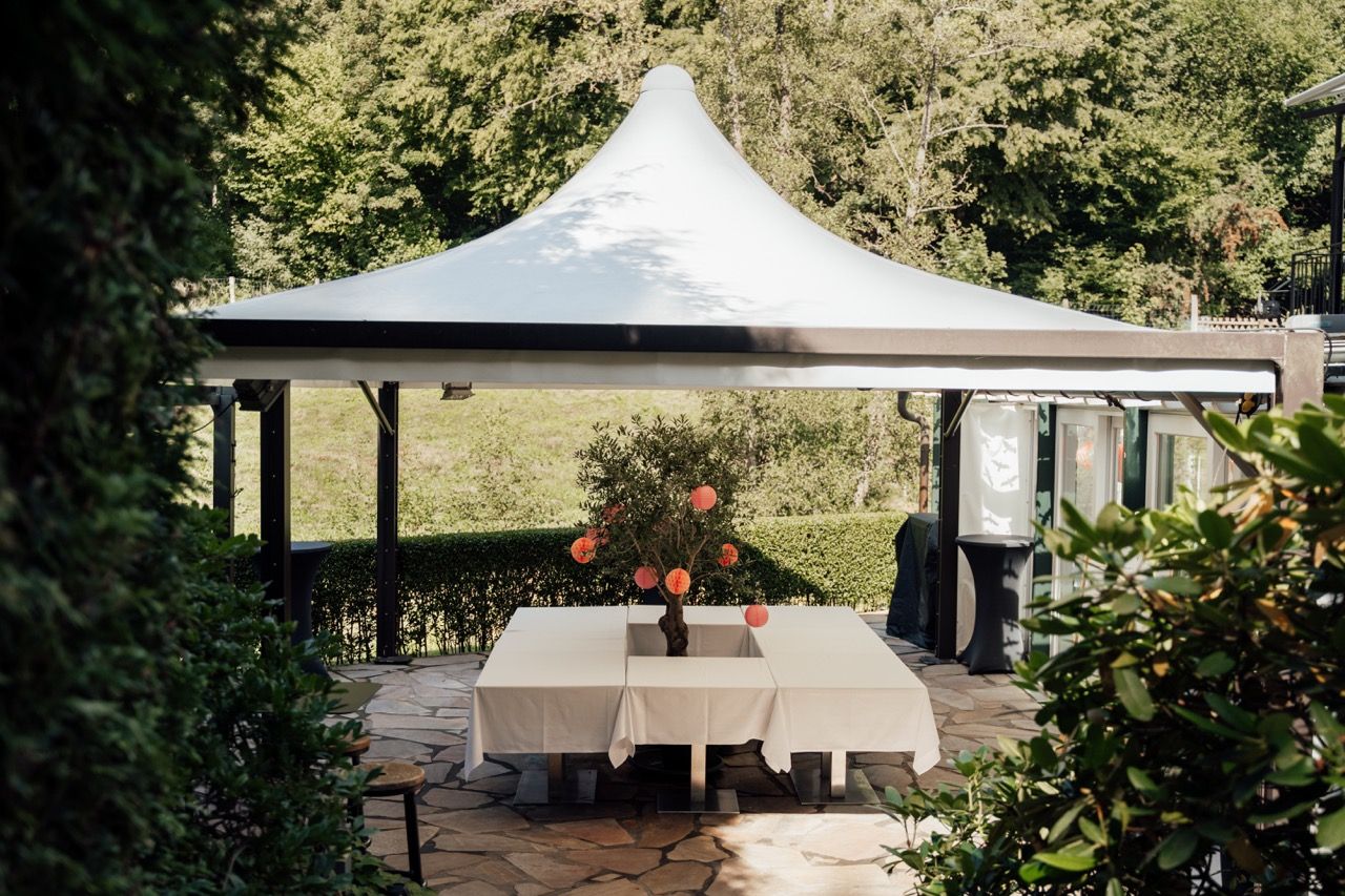 Elegant outdoor gazebo with white canopy over a table set with flowers on a stone patio surrounded by lush greenery.