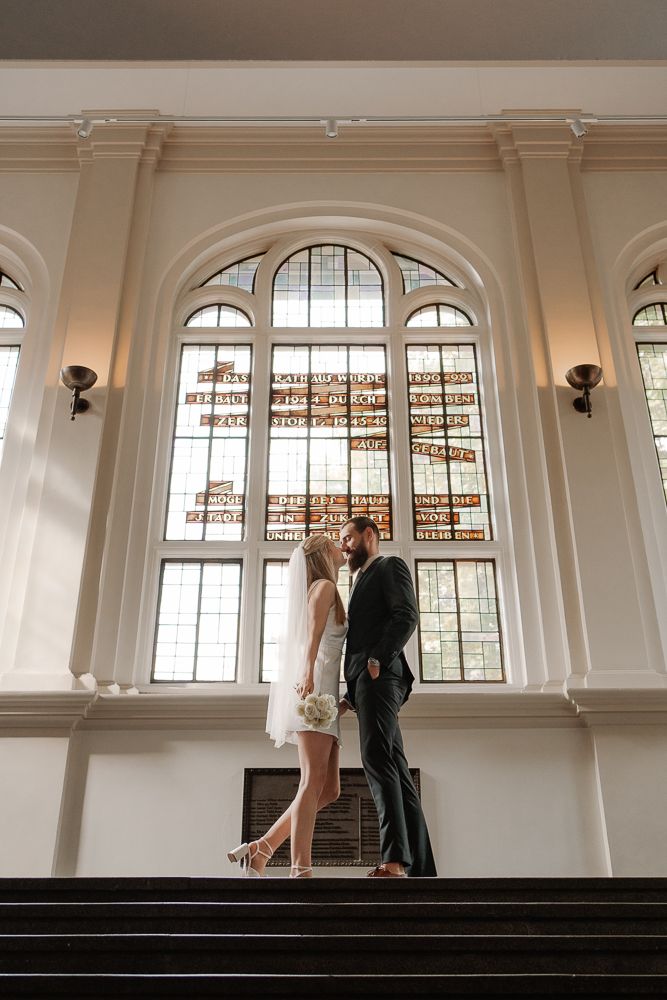 A couple stands together in an elegant white interior space with arched windows and high ceilings.