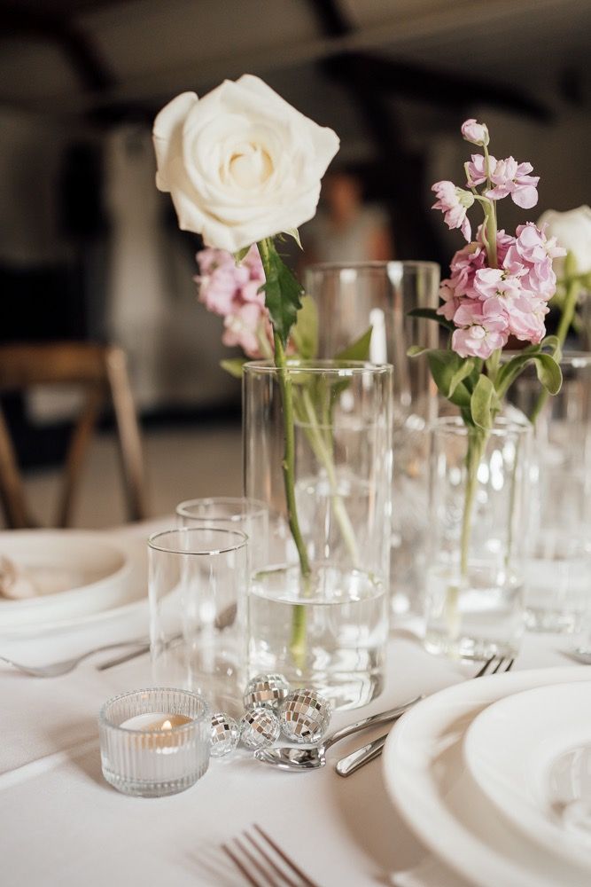 Elegant wedding table settings with delicate pink flowers and white roses in glass vases alongside place settings.