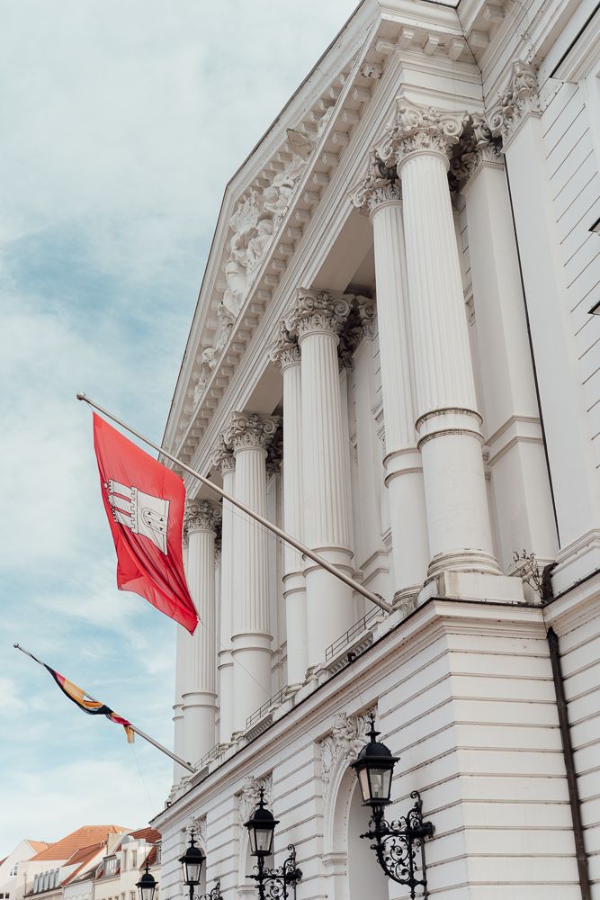 Ornate white neoclassical building facade with tall columns and red flags waving against a cloudy sky.