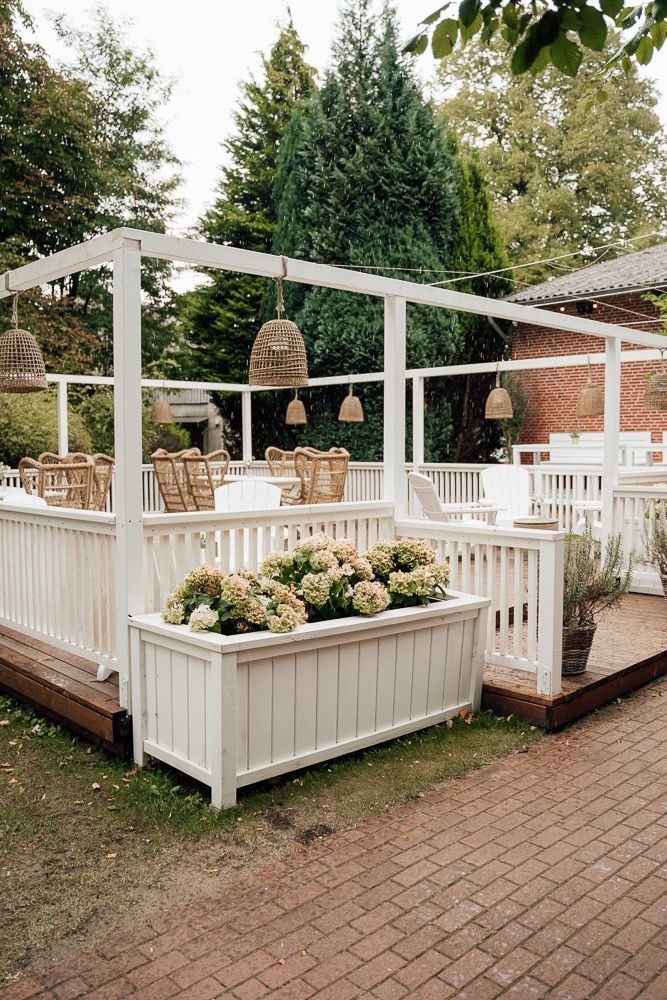 White wooden deck with railing and hanging wicker lanterns in an outdoor garden setting with brick paving.
