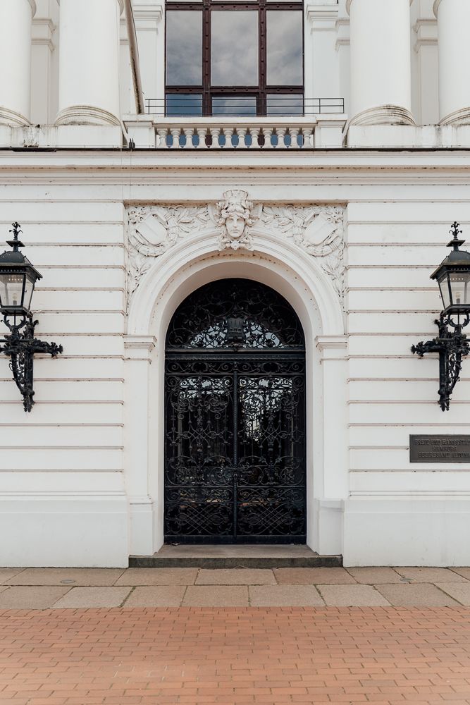 Elegant white building entrance with ornate black iron door, decorative archway and vintage wall-mounted lanterns.