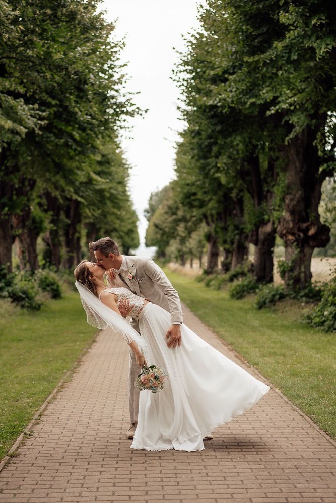 A romantic wedding couple dances and dips along a tree-lined brick pathway in an outdoor garden setting.