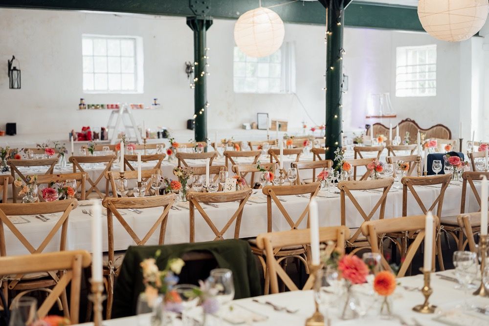 Long banquet tables with wooden cross-back chairs and white paper lanterns create an elegant reception setup in a bright venue.
