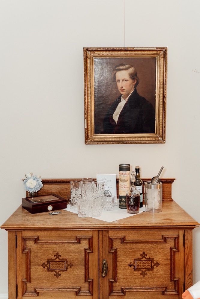 Vintage wooden dresser with ornate carvings displayed beneath framed portrait and decorative glassware in museum setting.