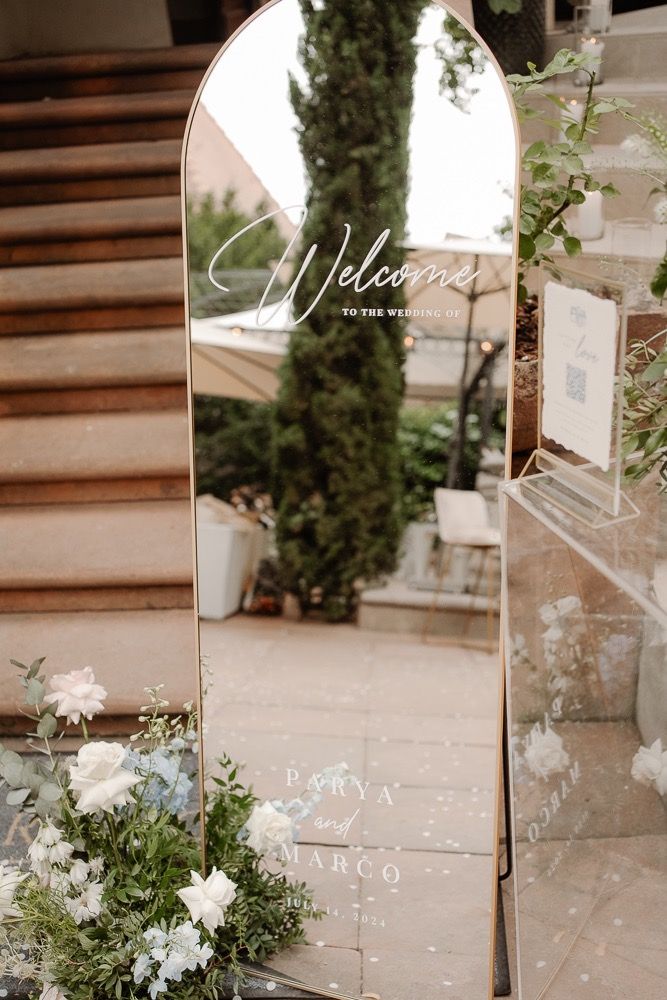 An elegant arched mirror with 'Welcome' text sits beside white floral arrangements and greenery at a wedding venue entrance.