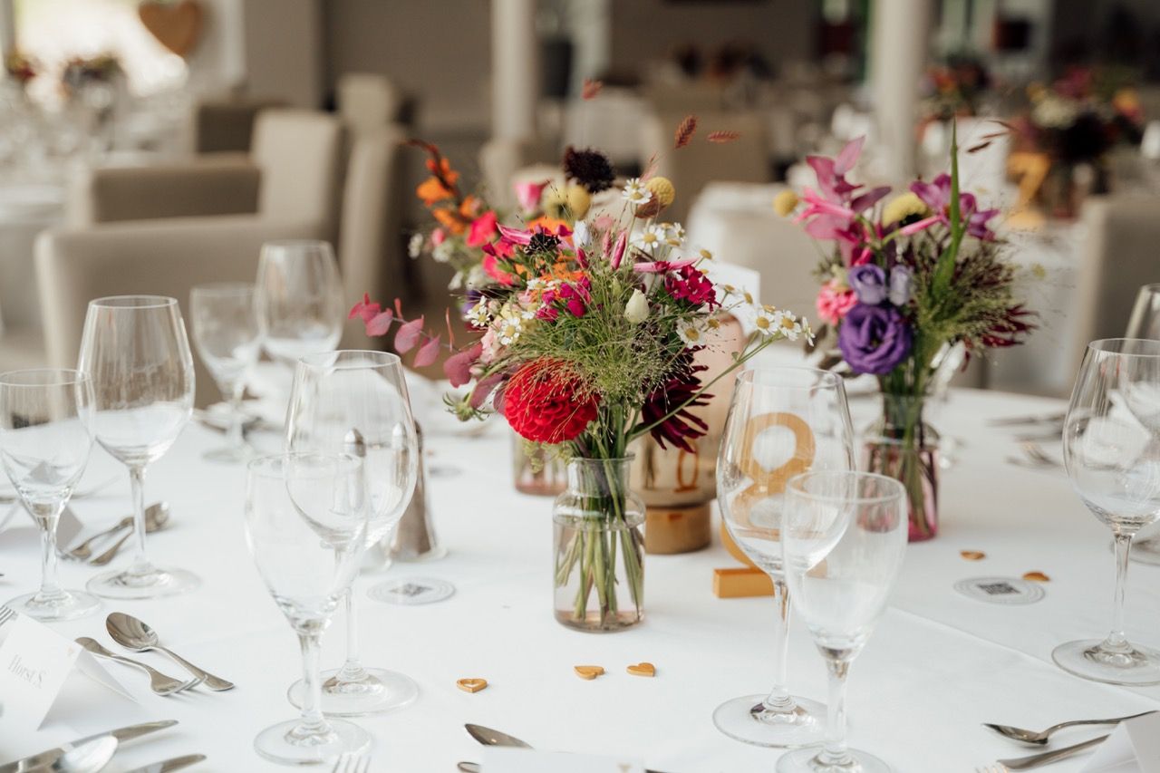 Elegant wedding reception table with white linen, crystal glasses, and colorful wildflower centerpieces in glass jars.
