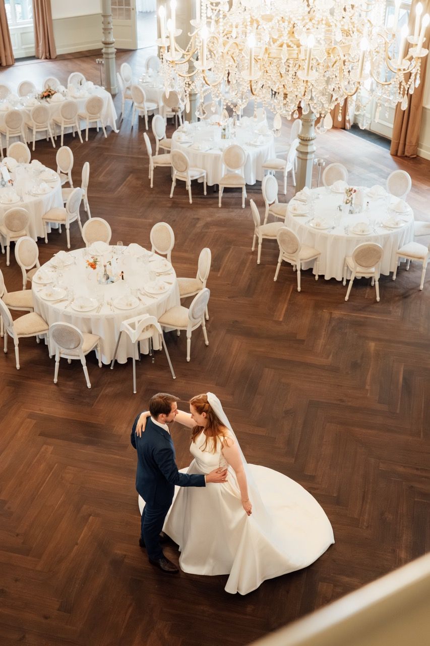 Bride and groom share their first dance in a grand ballroom with chandeliers and elegantly set round reception tables.