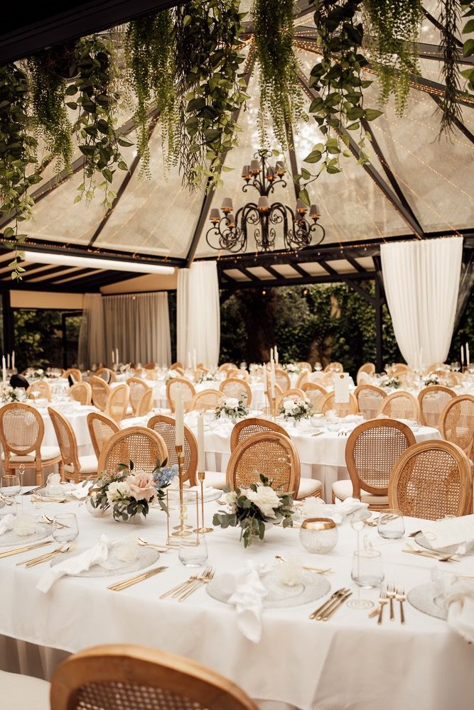 Elegant wedding reception tables set with white linens and rattan chairs in a greenhouse-style venue with natural lighting.