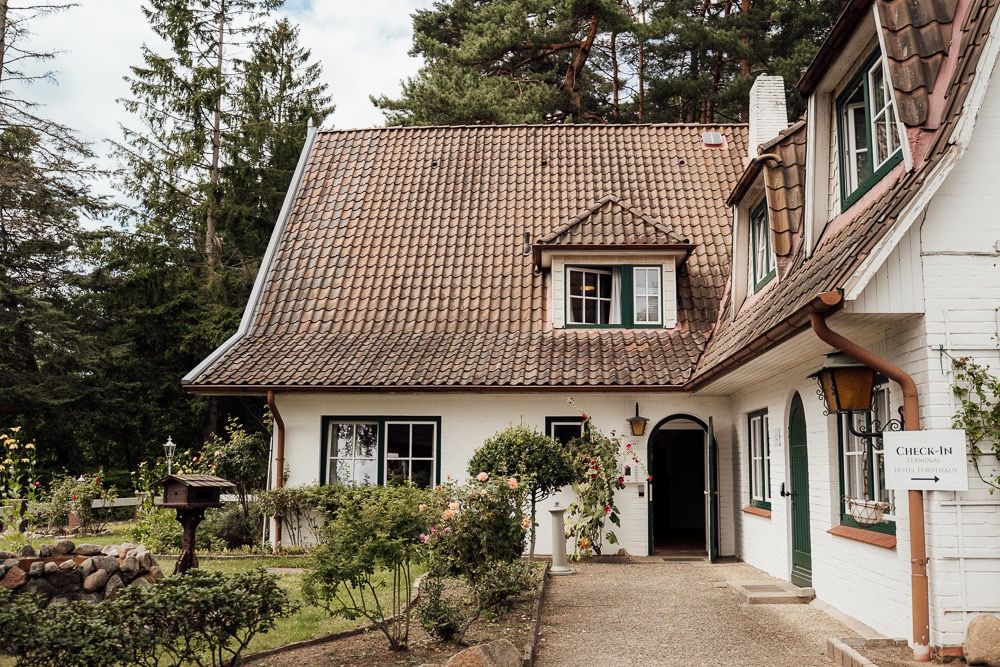 Charming white cottage with brown tiled roof and black shuttered windows nestled among trees in a forest setting.
