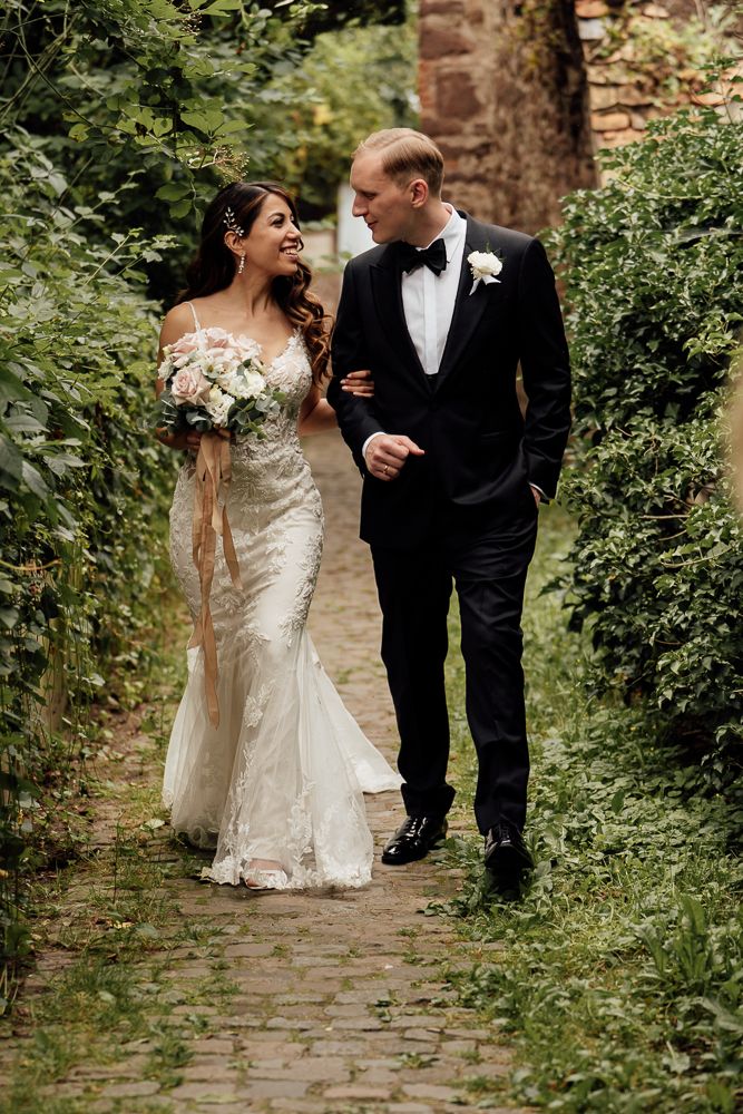A couple in formal wedding attire walks together on a cobblestone path surrounded by lush greenery.