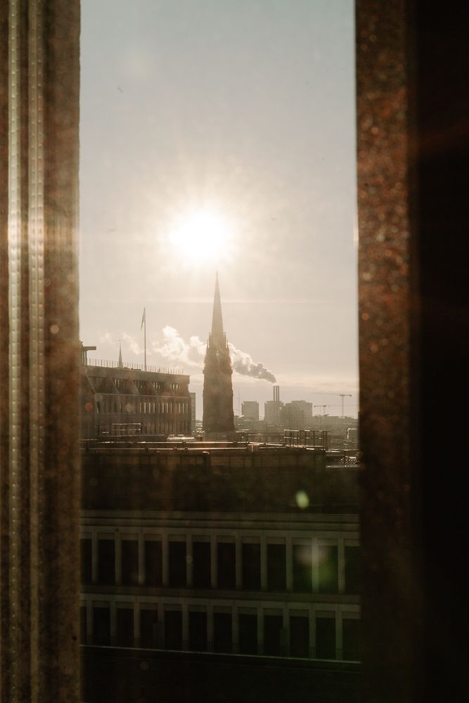 Moody urban cityscape viewed through window columns with industrial smoke stacks visible against overcast sky.