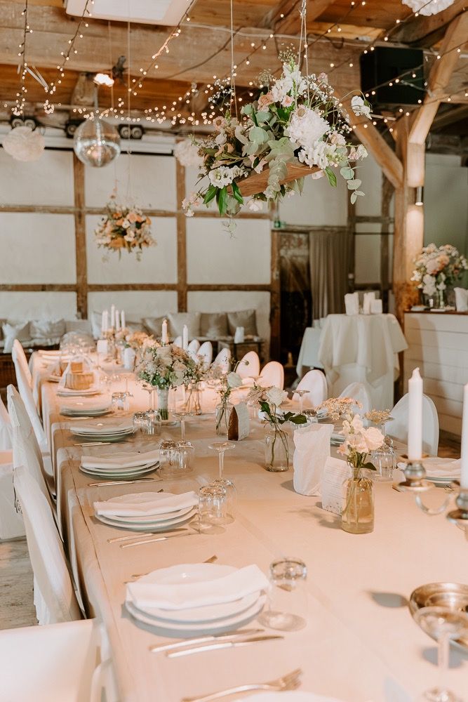 Elegant wedding reception tables set with white linens, soft pink accents, and rustic decor in a barn venue with twinkle lights.