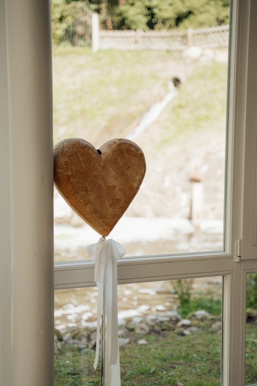 Wooden heart decoration with white ribbon bow hanging on a white window frame with garden view.