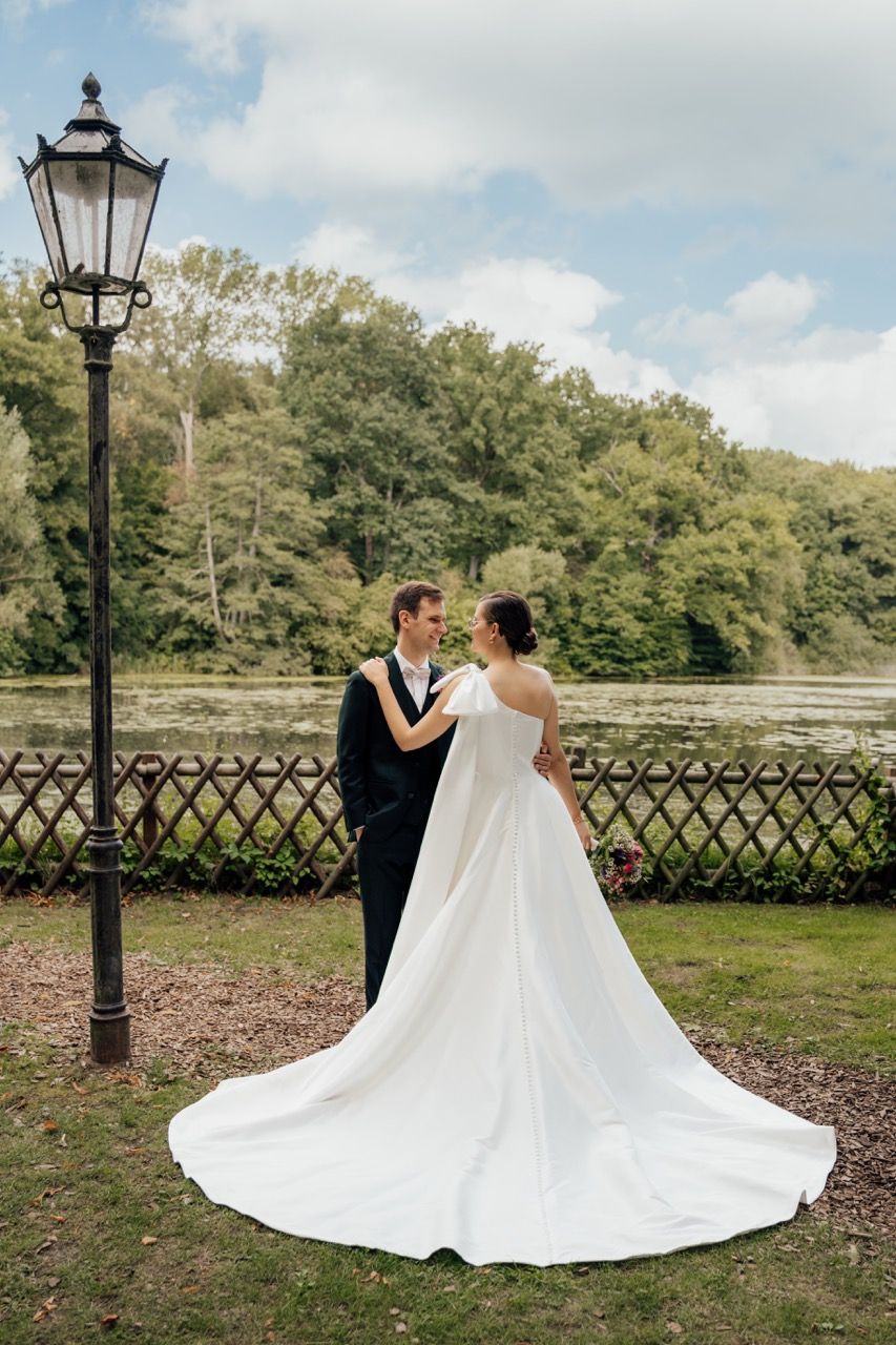 Bride in white gown with long train stands with groom in black tuxedo near vintage lamp post by serene lake.