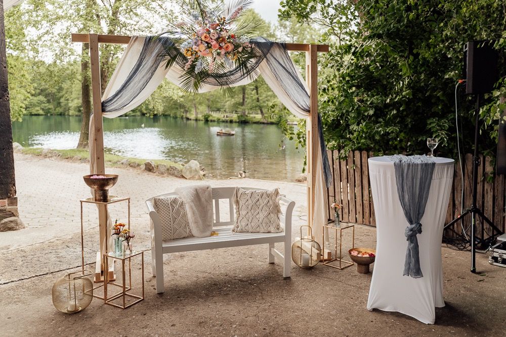 Rustic outdoor wedding venue setup with wooden pergolas, white drapes, and elegant decor along a lakeside setting.