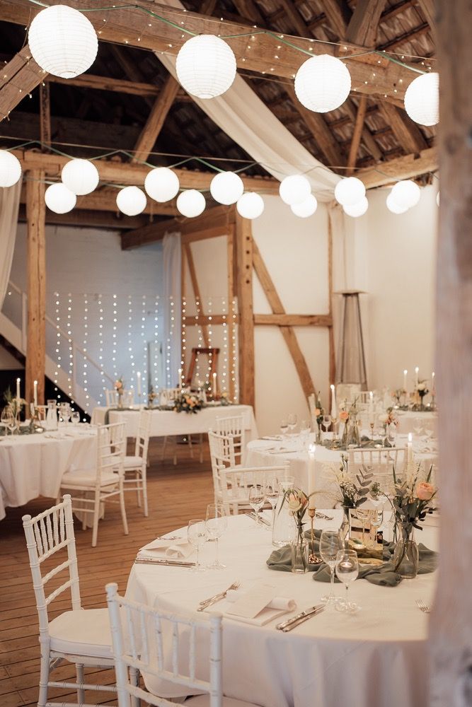 Rustic barn wedding reception venue with white paper lanterns, string lights, and elegant table settings on wooden floors.