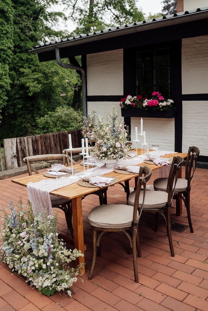 Rustic outdoor dining setup on brick patio with wooden table, cream cushioned chairs, and floral arrangements.