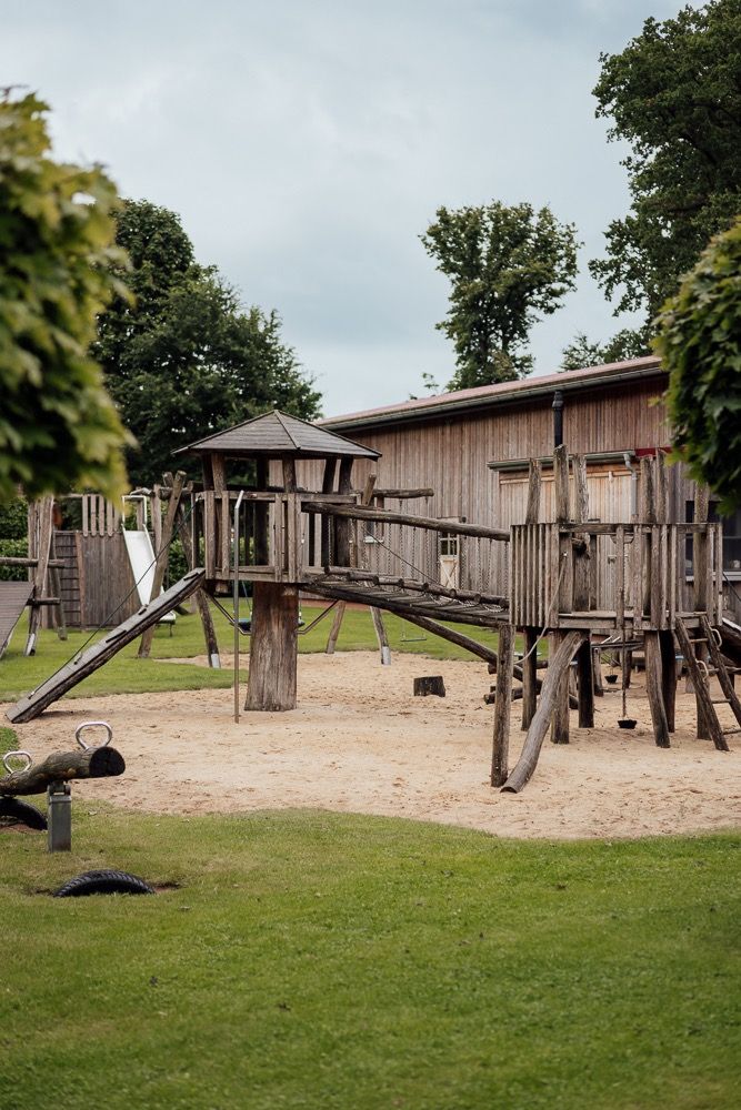 A rustic wooden playground structure with climbing elements and platforms sits on a sandy surface in a grassy park area.